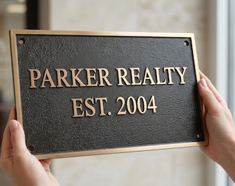 May include: A rectangular metal sign with a textured black finish and a smooth gold-toned border. The sign features raised gold-toned lettering that reads PARKER REALTY and EST. 2004. There are four small circular mounting holes in the corners.