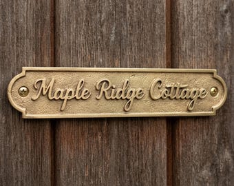 May include: A rectangular brass plaque with rounded ends and a textured surface, featuring the raised script text "Maple Ridge Cottage" in the center. The plaque is secured with two brass screws, one on each end, against a wooden surface.