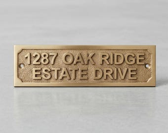 May include: A rectangular brass house sign with a textured background and raised lettering that reads 1287 OAK RIDGE ESTATE DRIVE. The metal plaque features a smooth border and two pre-drilled circular holes on the left and right sides.