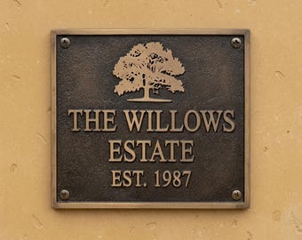 May include: A square dark bronze metal plaque with a textured finish, featuring a raised tree icon and the text THE WILLOWS ESTATE EST. 1987. The plaque is secured at each corner with a visible metal screw and has a smooth, raised border frame.