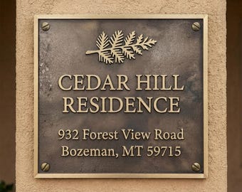 May include: A square bronze-toned metal plaque with a raised fern leaf icon at the top. The text reads CEDAR HILL RESIDENCE, 932 Forest View Road, Bozeman, MT 59715. The plaque is secured to a textured wall with four visible corner screws.
