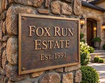 May include: A rectangular bronze-toned metal plaque mounted on a stone pillar. The plaque features raised lettering that reads "FOX RUN ESTATE" and "EST. 1991" in a serif font. The surface has a weathered, dark patina finish with four visible screws.