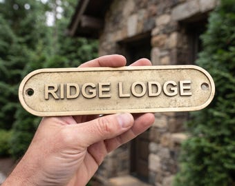 May include: An oval-shaped brass plaque with a weathered, antique finish featuring the raised, embossed text RIDGE LODGE in capital letters. The sign includes two circular mounting holes on the far left and right ends for secure installation.