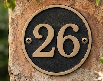May include: A circular house number plaque featuring the number 26 in raised gold-toned numerals against a textured black background. The plaque has a smooth gold-toned border and is secured with two visible screws on the left and right sides.