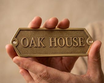 May include: A rectangular brass house sign with beveled edges and a textured surface. The sign features the embossed text OAK HOUSE in a serif font and includes two circular mounting holes on the left and right sides of the metal plate.