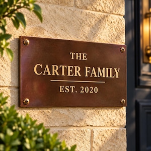 May include: A rectangular dark brown metal plaque with gold lettering reading THE CARTER FAMILY and EST. 2020. The sign features a horizontal line between the text and is secured to a light-colored stone wall with four visible metal screws.
