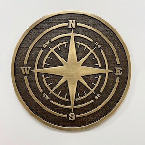 May include: A circular wall plaque featuring a raised brass-toned compass rose design against a dark, textured brown background. The compass displays the cardinal directions N, S, E, and W, along with the intermediate points nw, ne, sw, and se.