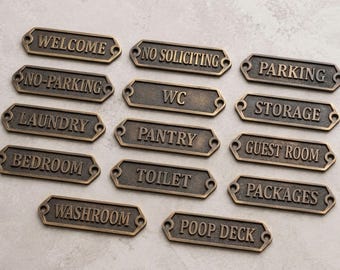 May include: A collection of fourteen rectangular bronze-toned metal signs with rounded ends and screw holes. Each sign features raised lettering including Welcome, No Soliciting, Parking, No-Parking, WC, Storage, Laundry, Pantry, Guest Room, Bedroom, Toilet, Packages, Washroom, and Poop Deck.