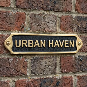 Puede incluir: Un letrero rectangular negro y dorado con las palabras "URBAN HAVEN" en letras doradas. El letrero está montado en una pared de ladrillos rojos. El letrero tiene un borde dorado y tornillos dorados en cada esquina.