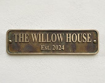 May include: A rectangular bronze-toned metal plaque with a weathered, dark patina finish. The sign features raised, polished lettering that reads THE WILLOW HOUSE and Est. 2024. It is secured by two brass-colored screws on the left and right sides.