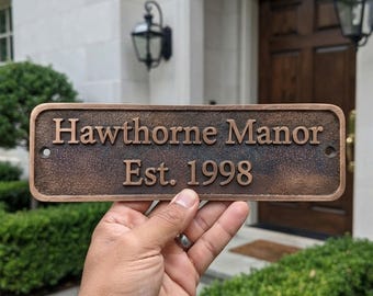 May include: A rectangular bronze-toned metal plaque with a textured, dark brown finish. The raised lettering reads "Hawthorne Manor" on the top line and "Est. 1998" on the bottom line. The plaque features rounded corners and two mounting holes.