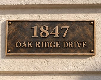 May include: A rectangular bronze-toned metal address plaque featuring the raised numbers 1847 and the text OAK RIDGE DRIVE in a serif font. The plaque has a textured, weathered finish and is secured at each corner with a small circular bolt.