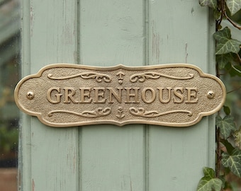 May include: A rectangular brass-colored metal sign with decorative scrollwork and small anchor icons. The word GREENHOUSE is embossed in capital letters across the center. The sign features a textured background and is secured with two screws.