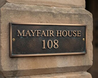 May include: A rectangular bronze-toned metal house plaque with a textured dark finish. The plaque features the raised text MAYFAIR HOUSE and the number 108 in a serif font. It is secured to a stone surface with two visible brass screws.