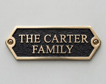 May include: A rectangular brass plaque with beveled edges and a textured black center. The plaque features the raised gold-colored text THE CARTER FAMILY in a serif font. It is secured by two visible screws on the left and right sides.