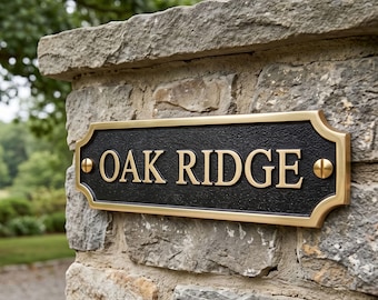 May include: A rectangular metal address plaque with a black textured background and a polished gold-colored border. The plaque features the raised gold-colored text OAK RIDGE and is secured to a stone surface with two gold-colored screws.