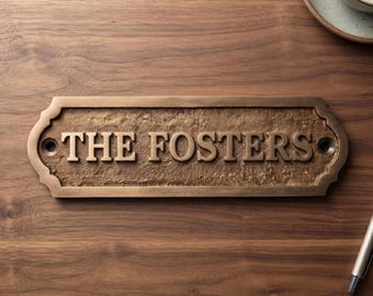 May include: A rectangular bronze-toned metal plaque with a textured, weathered surface and scalloped edges. The plaque features the raised, embossed text "THE FOSTERS" in a classic serif font, with a circular mounting hole on each side.