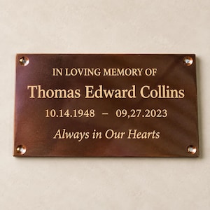 May include: A rectangular bronze memorial plaque with four corner screw holes. The engraved text reads: IN LOVING MEMORY OF Thomas Edward Collins 10.14.1948 &ndash; 09.27.2023 Always in Our Hearts. The metal surface has a polished, dark finish.