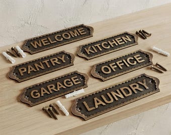 May include: A set of six rectangular metal signs with scalloped edges, featuring raised bronze-toned lettering on a dark textured background. The signs display the words WELCOME, KITCHEN, PANTRY, OFFICE, GARAGE, and LAUNDRY, accompanied by mounting screws and plastic wall anchors.