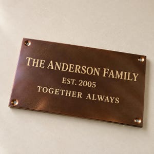 May include: A rectangular bronze-toned metal plaque featuring the engraved text THE ANDERSON FAMILY, EST. 2005, and TOGETHER ALWAYS. The plaque has a smooth, dark finish with four circular mounting holes, one located in each of the four corners.