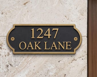 May include: A rectangular house address plaque with a black background and a gold-colored border. The plaque features the gold-colored text 1247 OAK LANE and is secured with two visible gold-colored screws on the left and right sides.