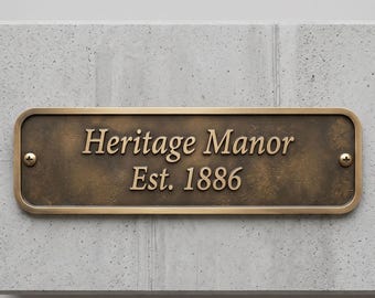 May include: A rectangular bronze-toned metal plaque with rounded corners and two visible screws. The plaque features the embossed text "Heritage Manor" on the top line and "Est. 1886" on the bottom line in a serif font.