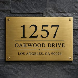 May include: A rectangular brushed brass house sign with four corner screws. The sign features the black engraved text 1257, OAKWOOD DRIVE, and LOS ANGELES, CA 90026, separated by horizontal lines and a small centered dot.