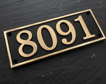 May include: A rectangular house number plaque featuring the digits 8091 in a metallic gold finish. The numbers are set against a matte black background and framed by a gold border, with four small circular mounting holes at each corner.