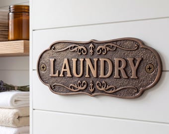May include: An oval-shaped bronze-colored metal wall plaque with a textured background and raised lettering that reads LAUNDRY. The plaque features decorative scrollwork and small anchor motifs, secured to a white wall with two visible screws.
