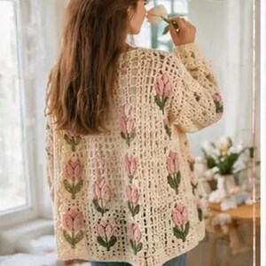 May include: Cream-colored, open-weave crochet cardigan with pink tulip floral accents. The long-sleeved sweater features a delicate, airy design, perfect for layering. The floral pattern is repeated across the front and sleeves.