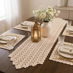 Puede incluir: Una mesa de comedor puesta con un camino de mesa de ganchillo color crema y manteles individuales a juego. Una linterna dorada con una vela encendida se encuentra sobre el camino de mesa, junto a un jarrón blanco con flores y cubiertos.