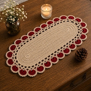 May include: A cream-colored, oval crochet table runner with a scalloped edge of red floral designs. The runner is placed on a dark wooden surface, with a vase of white flowers, a lit candle, and a pinecone nearby.
