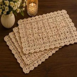 May include: Four beige crochet placemats with a floral design and scalloped edges are stacked on a dark wooden surface. A white vase with flowers and a lit candle in a glass holder are in the background.