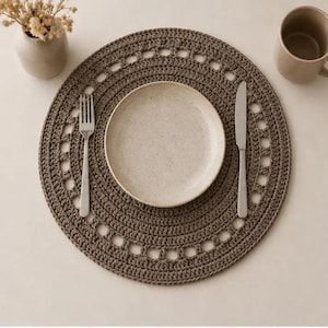 Puede incluir: Un mantel individual gris tejido a ganchillo con un borde calado decorativo, un plato blanco, un tenedor y un cuchillo. También hay una taza beige y un pequeño jarrón con flores secas, lo que sugiere un entorno de comedor.