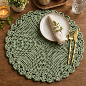 May include: A sage green, crocheted placemat with a scalloped edge. A white plate with a rolled napkin and sprig of greenery sits atop the placemat. Gold-colored fork and knife are placed to the right. The placemat is on a wooden table.