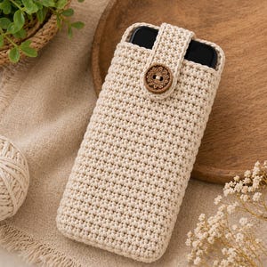 May include: Cream-colored, crocheted phone case with a wooden button closure. The case is rectangular and has a textured surface. The button is round with a floral design. The phone case is designed to hold a smartphone.