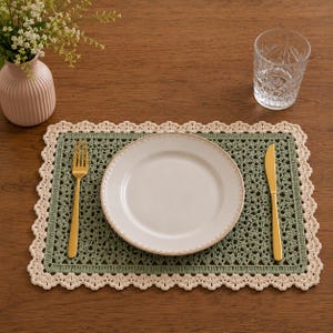May include: A rectangular crochet placemat featuring a sage green center with a decorative cream-colored scalloped border. A white ceramic plate sits in the center, flanked by a gold-toned metal fork on the left and a gold-toned knife on the right.