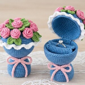 May include: Two blue crochet ring boxes, one open with a ring inside. Each box has a pink bow and is topped with pink crochet roses and green leaves. The open box has a white scalloped edge.