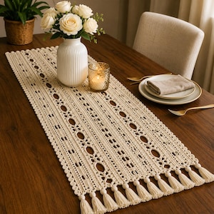 Peut inclure: Chemin de table crème en tissu ajouré avec un motif décoratif et des glands. Il est posé sur une table en bois foncé, avec un vase blanc rempli de roses, une bougie et des couverts.