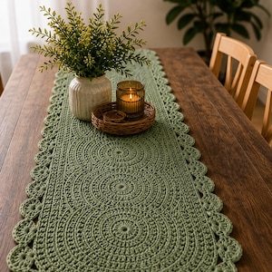 Peut inclure: Chemin de table en crochet vert sauge avec bordure festonnée et motif circulaire. Un vase avec de la verdure, une bougie dans un photophore et un plateau en osier sont posés sur une table en bois foncé. La table est dressée pour un repas rustique et chaleureux.
