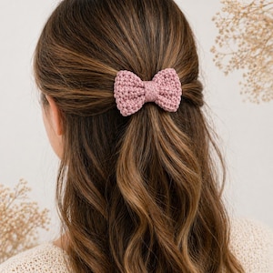 May include: A dusty rose crochet bow hair clip. The bow is centered in the hair, with the wearer's back to the camera. The hair is a mix of brown and blonde tones. The bow is made of a textured yarn.