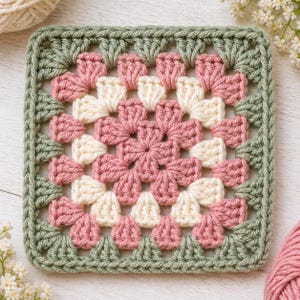 May include: A square, handmade crochet piece in a granny square pattern. The design features a central flower pattern in pink and cream, surrounded by a border of green. The yarn appears to be soft and textured, perfect for blankets or other crafts.