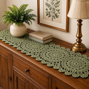 Peut inclure: Un chemin de table en crochet vert sauge présentant un motif circulaire répétitif de type mandala avec des bords festonnés. Le chemin est réalisé en fil avec des détails ajourés complexes, montrant une série de motifs floraux reliés.