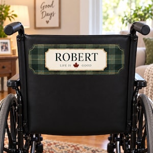 Peut inclure: Un fauteuil roulant noir avec une plaque rectangulaire fixée sur le dossier. La plaque présente une bordure à carreaux vert foncé et un centre crème avec le texte "ROBERT" et "LIFE IS GOOD" séparés par un petit motif de feuille d'érable rouge.