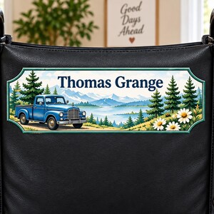 Peut inclure: Une plaque rectangulaire fixée au dossier d'un fauteuil roulant noir. L'illustration représente un pick-up bleu, des sapins, des montagnes, un lac et des marguerites blanches, avec le texte Thomas Grange inscrit en lettres bleues.