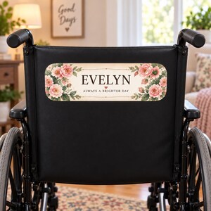 Peut inclure: Un fauteuil roulant noir avec une plaque rectangulaire fixée sur le dossier. La plaque présente un fond crème avec des illustrations florales roses sur les côtés, le nom EVELYN en noir et la phrase ALWAYS A BRIGHTER DAY inscrite en dessous.