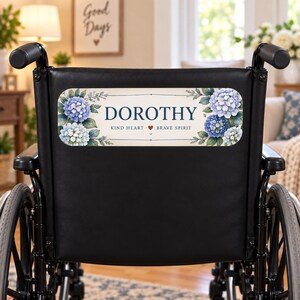 Peut inclure: Un fauteuil roulant noir avec une plaque rectangulaire fixée au dossier. La plaque affiche le nom DOROTHY en bleu foncé, suivi de KIND HEART, une petite icône de cœur rouge et BRAVE SPIRIT, le tout encadré par des fleurs d'hortensia bleues.