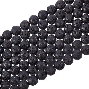 Peut inclure: Un long cordon de perles de lave noires et rondes disposées en diagonale. Chaque perle présente une surface poreuse et texturée caractéristique de la roche volcanique, créant un aspect uniforme sur toute la longueur du fil.