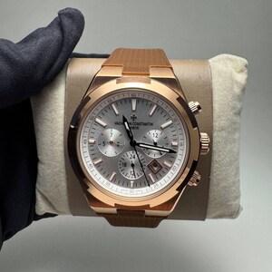 Op de afbeelding: Een chronograaf polshorloge van roségoud met een zilveren wijzerplaat met drie sub-wijzerplaten, een datumvenster en de tekst Vacheron Constantin Geneve. Het horloge heeft een bruine, gestructureerde rubberen band en een gepolijste, gekartelde lunette.