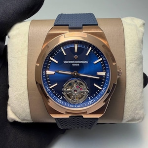 Peut inclure: Une montre-bracelet en or rose avec un cadran bleu et un bracelet en caoutchouc bleu. Le cadran de la montre porte la marque "VACHERON CONSTANTIN GENEVE". La montre est présentée sur un coussin beige.